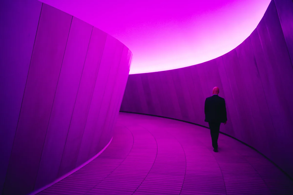 Figure walking through a curved concrete corridor lit in magenta