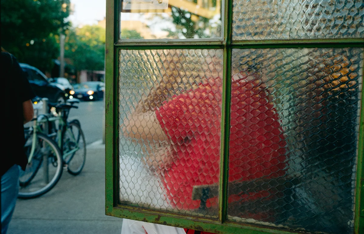 Person in a red jacket seen through textured glass in a green window frame, with bicycles and a street behind